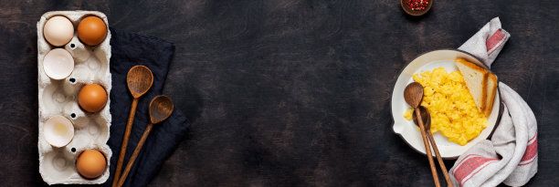 炒鸡蛋和生鸡蛋在盒子里，餐巾和木汤匙，深色木桌子的背景。传统的英式早餐。前视图。模拟。横幅。图片下载