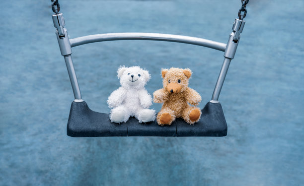 两个微笑的泰迪熊坐在操场的秋千上，快乐的棕色和可爱的白熊坐在一起在阳光明媚的春天或夏天的公园。素材图片