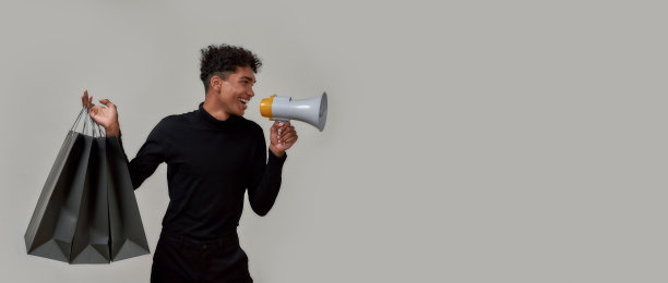 酷酷的年轻人穿着优雅的黑色在扩音器中尖叫，做销售公告，与购物袋在灰色的背景孤立图片下载