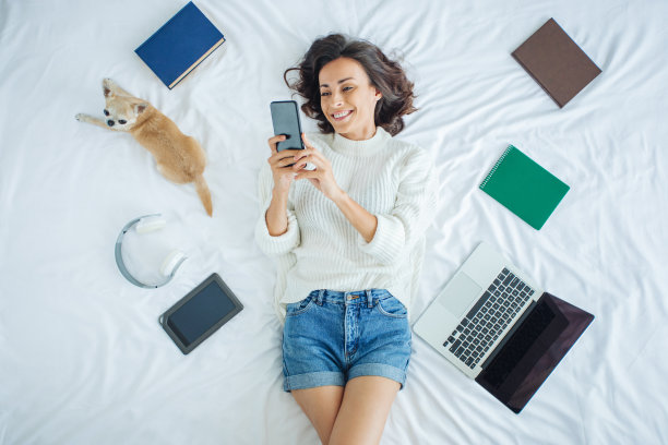 俯视图照片，一个迷人的穿着休闲微笑的女孩躺在床上和她可爱的吉娃娃狗在浏览智能手机上的社交媒体应用程序图片下载