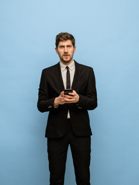 英俊的年轻人在黑色西装与电话孤立在蓝色背景。图片下载