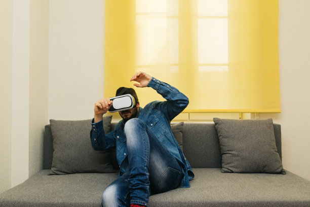 一个戴着眼镜和虚拟现实头戴式耳机的吓坏了的男人躺在家里的沙发上。3d家居的创新概念。图片下载