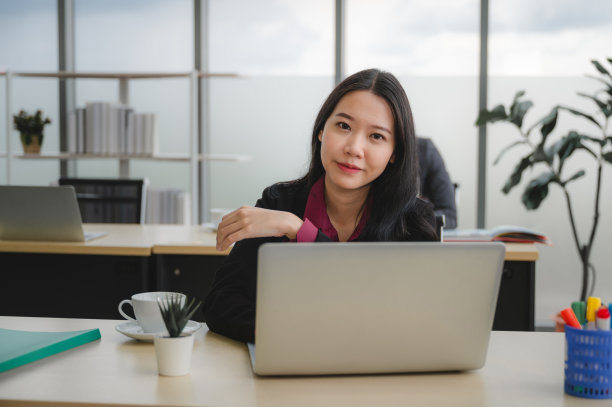 自信和聪明美丽的女商人在正式的着装站在办公室。背景是一群朋友和笔记本电脑。在工作场所工作是一个令人愉快的想法。图片下载