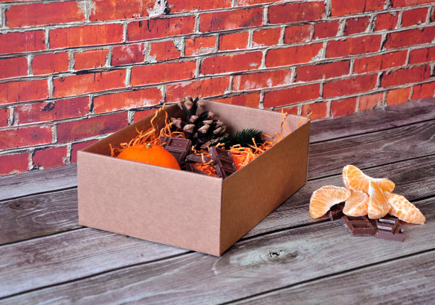 靠砖墙的木桌上放着一个纸板箱，里面放着橘子、巧克力和松果。图片下载