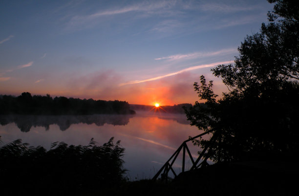 雾蒙蒙的湖面上，太阳和树木的倒影呈现出橘黄色的日出。清晨清新的风景。水面上的薄雾，森林的轮廓和初升的太阳的光线。图片下载