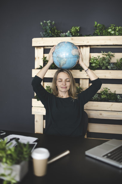 年轻漂亮的金发美女带着地球仪坐在笔记本电脑前，在一间非常现代、生态、可持续的办公室里梦想着度假图片下载