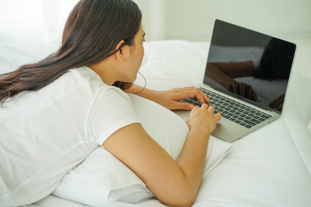 亚洲年轻漂亮的女人从家里工作与笔记本电脑在更白的床上图片下载
