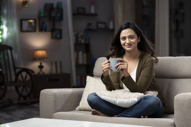 女人拿着咖啡杯，坐在家里的沙发上喝咖啡:-库存照片图片下载