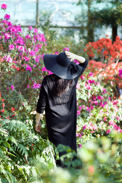 有选择性的重点。一个女孩穿着优雅的黑色连衣裙和帽子在盛开的杜鹃花花园里。从后面看。春季概念，3月8日放假图片下载