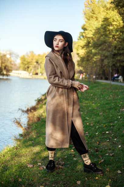 时尚的年轻女士在长米色外套和黑帽子走在城市绿色公园的河边图片下载