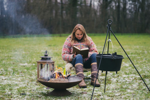 一个穿着羊毛衫的金发女郎在篝火旁的草地上看书。火碗里烧着火，上面挂着一只烧开的水壶。在冬天的傍晚，日落时分露营图片下载