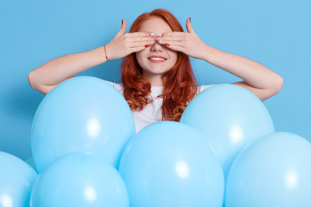 年轻女子与许多蓝色气球在孤立的颜色背景，用手遮住眼睛和微笑，有惊喜在她的生日，欧洲女士表达积极的情绪在假日。图片下载