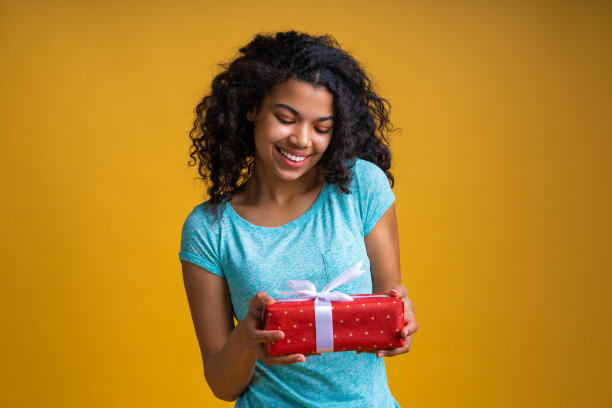 肖像美丽的穿着随意的黑皮肤女孩与完美的微笑拿着礼品盒装饰缎带在手里图片下载