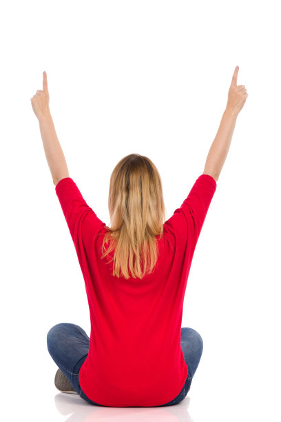 一名年轻女子坐在地板上，双臂举起并指向上方。后面的观点。图片下载
