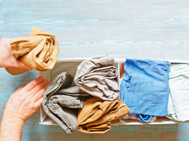 男士的手将卷起的裤子(裤子、牛仔裤)从箱子里拿出来或放进箱子里。整理男士衣柜的概念。附近的抽屉里还有其他衣服。木制背景。图片下载