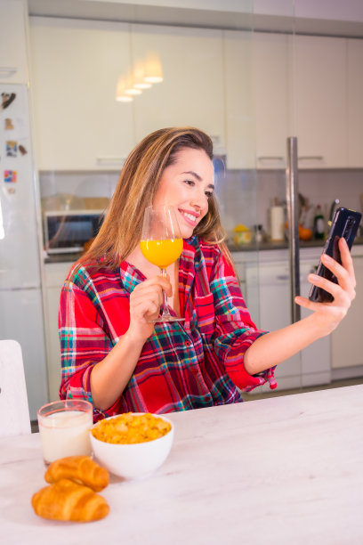 健康的生活方式，漂亮的金发白种女人喝橙汁当早餐，用手机自拍图片下载
