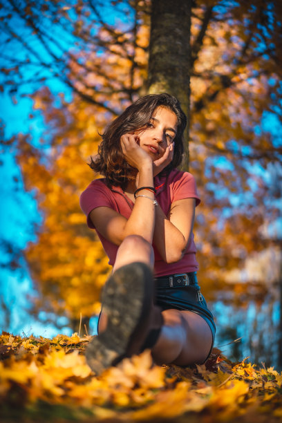 秋天的拍摄中，一个漂亮的黑发女孩穿着粉红色的衬衫坐在树下。自信的女孩的脸图片下载