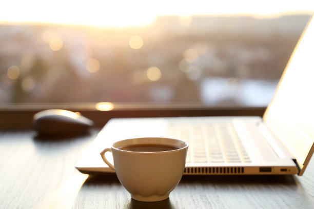舒适的工作场所在家庭办公室，热咖啡杯和笔记本电脑在桌子上对着窗户在日落图片下载