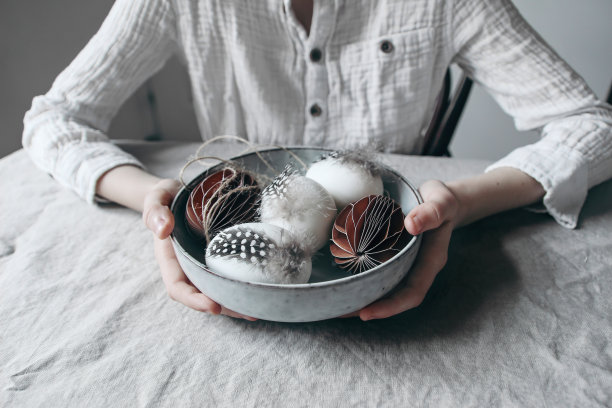 特写儿童的手在白色棉质衬衫拿陶瓷碗装饰复活节彩蛋和纸装饰。穆迪春生活作文。模糊的背景，选择性聚焦。图片下载