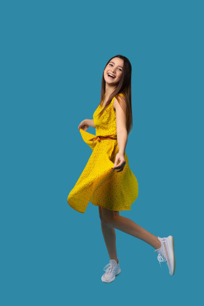 快乐的白人女孩在明亮的黄色连衣裙微笑和摆姿势的蓝色背景与侧空间。图片下载