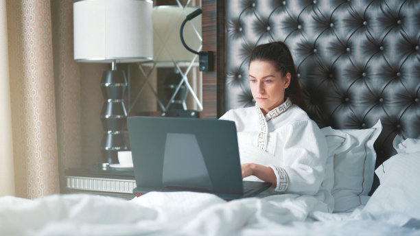 一个穿着白色浴衣的漂亮的黑发女子躺在床上，用笔记本电脑工作。公寓或酒店房间的漂亮内饰。美丽的早晨，新的一天。日常生活，女商人，成功图片下载