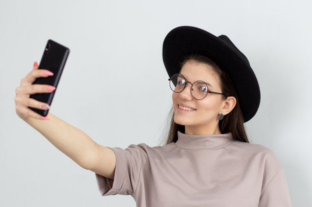 美丽的年轻女性与蓝色的眼睛拿着手机，摆姿势自拍，看着相机与微笑。有魅力的女人打视频电话。图片下载