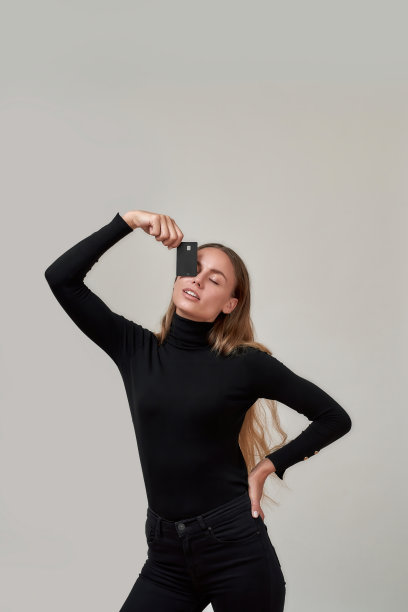 好玩的年轻女人穿着黑色的衣服看起来内容，拿着塑料信用卡在她的眼睛，而摆出孤立的灰色背景图片下载