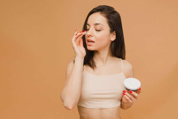 照片中迷人的年轻女子在内衣，把营养霜在她的皮肤上，孤立的米色背景。运动、健身、饮食、营养保健理念。图片下载