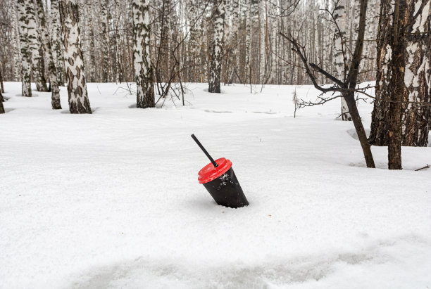 在冬天的森林里，一个带红色塑料盖的黑色咖啡杯从雪地里伸出来。生活垃圾对环境的污染图片下载