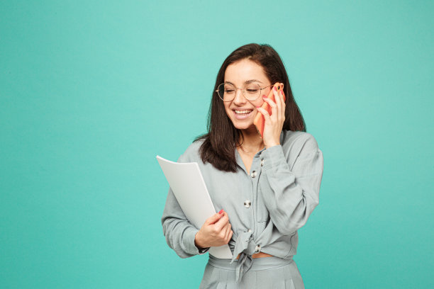 照片可爱的商务女士在眼镜持有文件和电话。身穿灰色衬衫，孤立的绿松石色背景图片下载