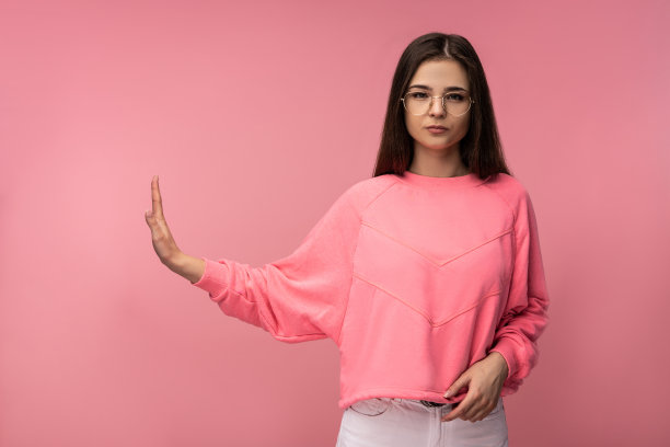 照片中的年轻美女戴着眼镜，严肃的脸和显示拒绝。穿休闲粉色t恤白色裤子孤立粉色背景图片下载