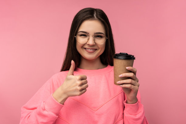 有魅力的女人在眼镜的照片持有咖啡和拇指向上。穿休闲粉色t恤白色裤子孤立粉色背景图片下载
