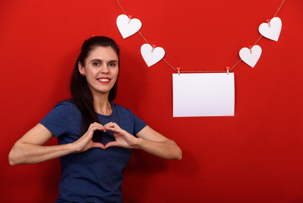 美丽的黑发女人穿着休闲的蓝色t恤在孤立的红色背景微笑近矩形旗帜和白色的心，显示心从手指图片下载