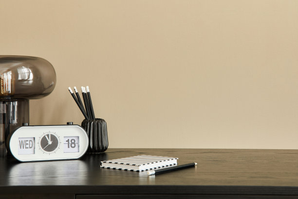 黑色书桌、设计台灯、白色时钟、笔记本电脑和办公桌管理器构成了办公室内部的时尚组合。模板。米色墙。副本的空间。图片下载