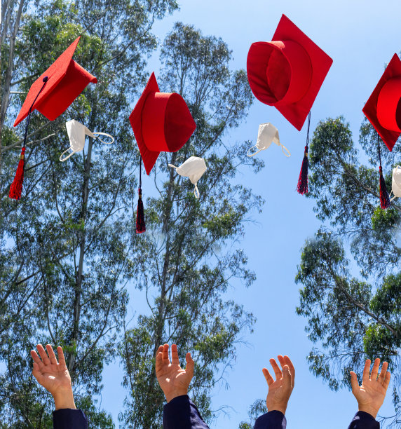 蓝色的天空上，学生们把红色的毕业帽和面具抛向空中。毕业和社交距离的概念图片下载