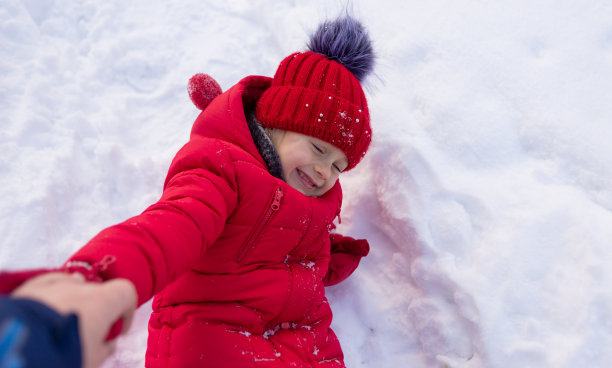 一个穿着红色夹克，戴着针织帽子的小女孩躺在洁白的雪地上，爸爸向她伸出手来。冬天孩子们的娱乐活动。图片下载