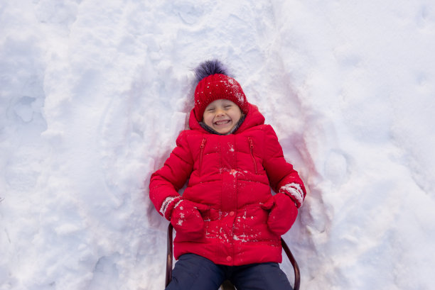 一个穿着红色夹克，戴着针织帽子的小女孩躺在洁白的雪地上。冬天孩子们的娱乐活动。图片下载
