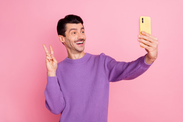 照片积极有趣的年轻人穿着紫色套头衫显示v标志钉自拍照现代设备孤立的粉红色背景图片下载