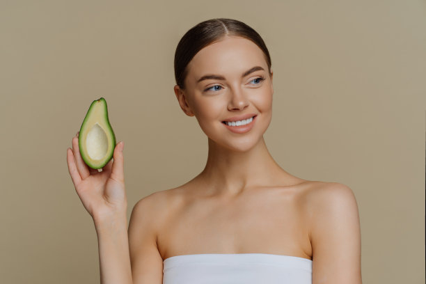 美容护肤理念。积极健康愉快的女人看起来温柔的一边微笑，温柔地拿着一半鳄梨，完美的化妆，雪白的牙齿包裹在浴巾上，孤立在棕色的工作室墙壁上图片下载