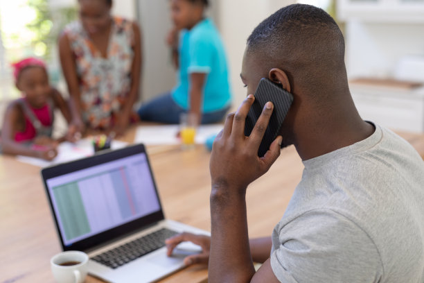 非裔美国父亲使用笔记本电脑和智能手机在家工作，有家庭背景图片下载