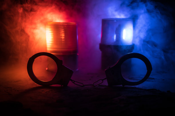 警方夜间突袭，你将被逮捕。手铐的轮廓和警车在后面。在雾蒙蒙的背景下，红色和蓝色警灯闪烁。图片下载