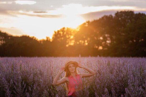 一个年轻女子的肖像，身穿洋红色衬衫，牛仔短裤，头上戴着草帽，背景是盛开的粉红色鼠尾草。夕阳下的金色长发。出城旅行图片下载