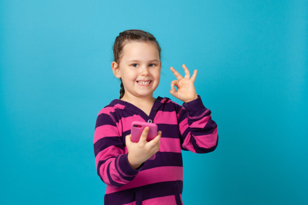 一个梳着辫子的微笑女孩的肖像，拿着一个粉红色的t型盒子里的手机，在蓝色背景上显示孤立的戒指手势图片下载