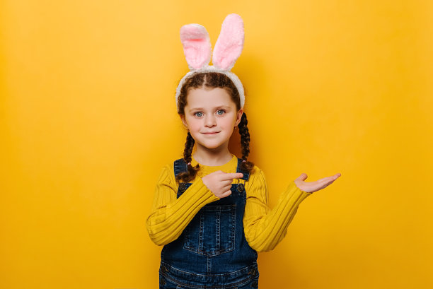 水平的照片，快乐的学龄前女孩在粉红色兔子毛茸茸的耳朵，指向一边的手指，展示了一些空白空间，孤立在黄色的工作室背景。复活节假期的概念图片下载