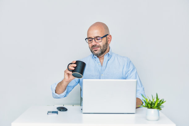 迷人的休闲男人戴着眼镜坐在办公桌前，在笔记本电脑上工作，同时举起一杯咖啡在你的家快乐。高质量的照片图片下载