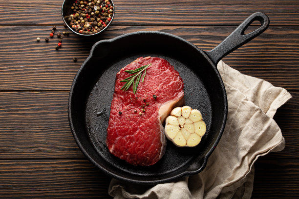新鲜的生牛肉配上调味料，大蒜和迷迭香，放在铸铁煎锅上，在黑色的木头背景上，上面用餐巾图片下载