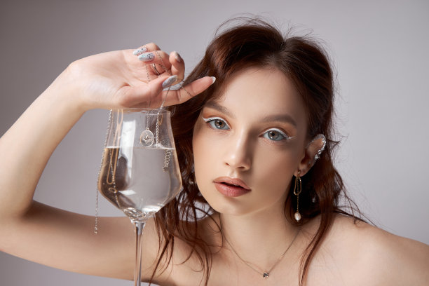 漂亮的年轻女子，手里拿着玻璃杯和珠宝。专业的妆容，富有表情的眼睛图片下载
