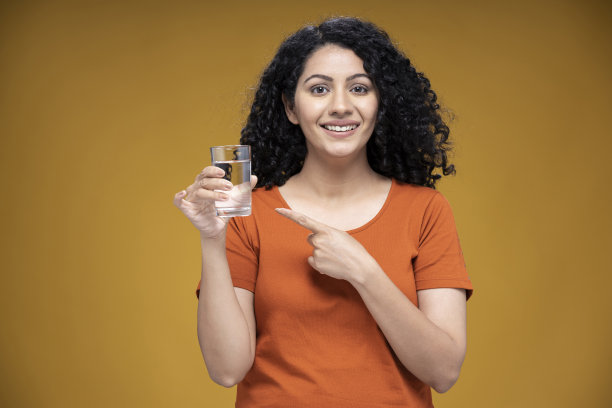 在黄色背景上，一个年轻的女人拿着并指着一杯纯净水:-库存照片图片下载