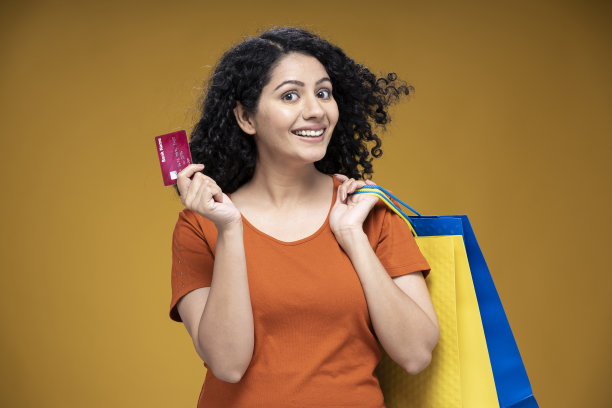 年轻女子的肖像提着购物袋，并显示信用卡站在孤立的黄色背景:-库存照片图片下载