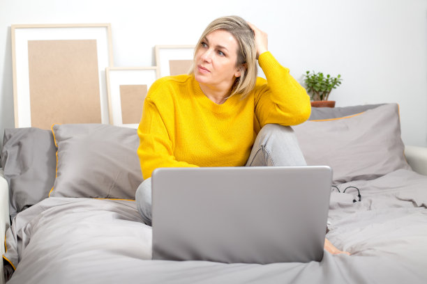 一个专注沉思的女人正坐在床上，一边用笔记本电脑工作，一边看着窗外，思考着一个策略。解决这个问题。在家工作，自由职业者。色彩趋势2021年图片下载